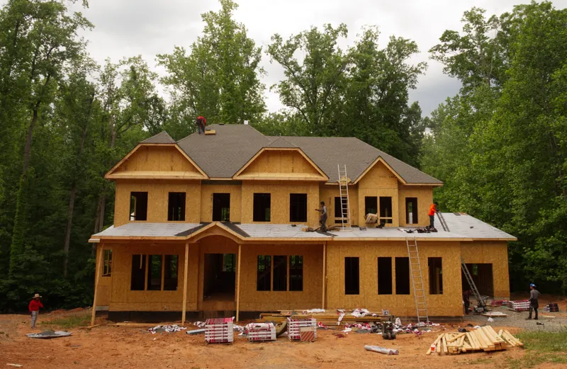 Front of a framed two-story house with the roof sheathing on, framing lumber staged in the yard