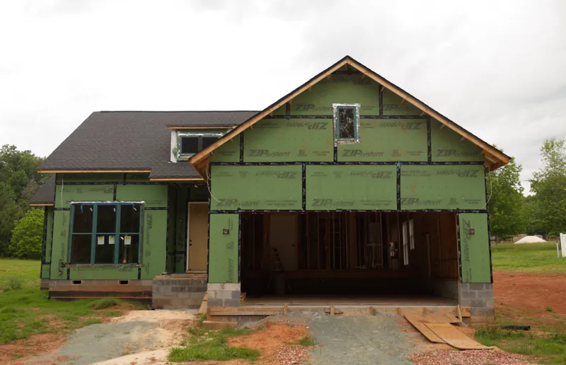 Single-story house with a front-facing gable and attached garage, wrapped in green structural sheathing before siding